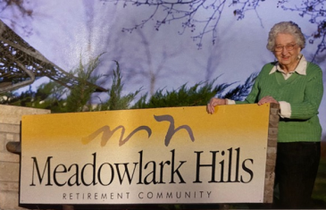 File photo. The late Margaret Wheat pictured with an sign that marked the entrance of Meadowlark’s campus from Kimball Avenue. Margaret joined the Meadowlark Hills board in 1976 and is credited for suggesting the community’s name.